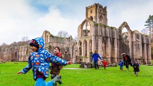 A family running across an open green space, with dramatic stone ruins behind